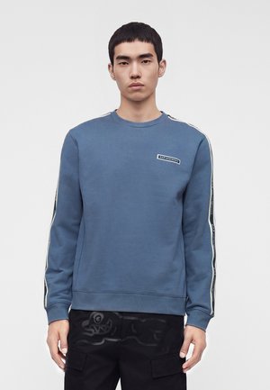 Jeune homme portant un sweat-shirt bleu à manches longues avec des bandes de marque sur les manches et un pantalon noir à poches cargo, debout devant un fond simple.