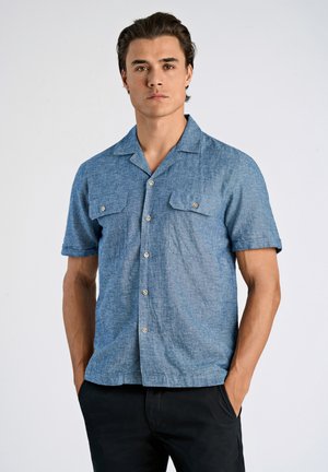 Camisa de chambray azul de manga corta con botones en la parte delantera, dos bolsillos en el pecho y un cuello de tipo camp. La tela tiene un aspecto texturizado.