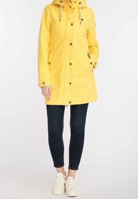 Gul vattentät jacka med huva, framknappning, dragsko och två sidofickor. Bärs med mörka slim fit-jeans och vita sneakers.