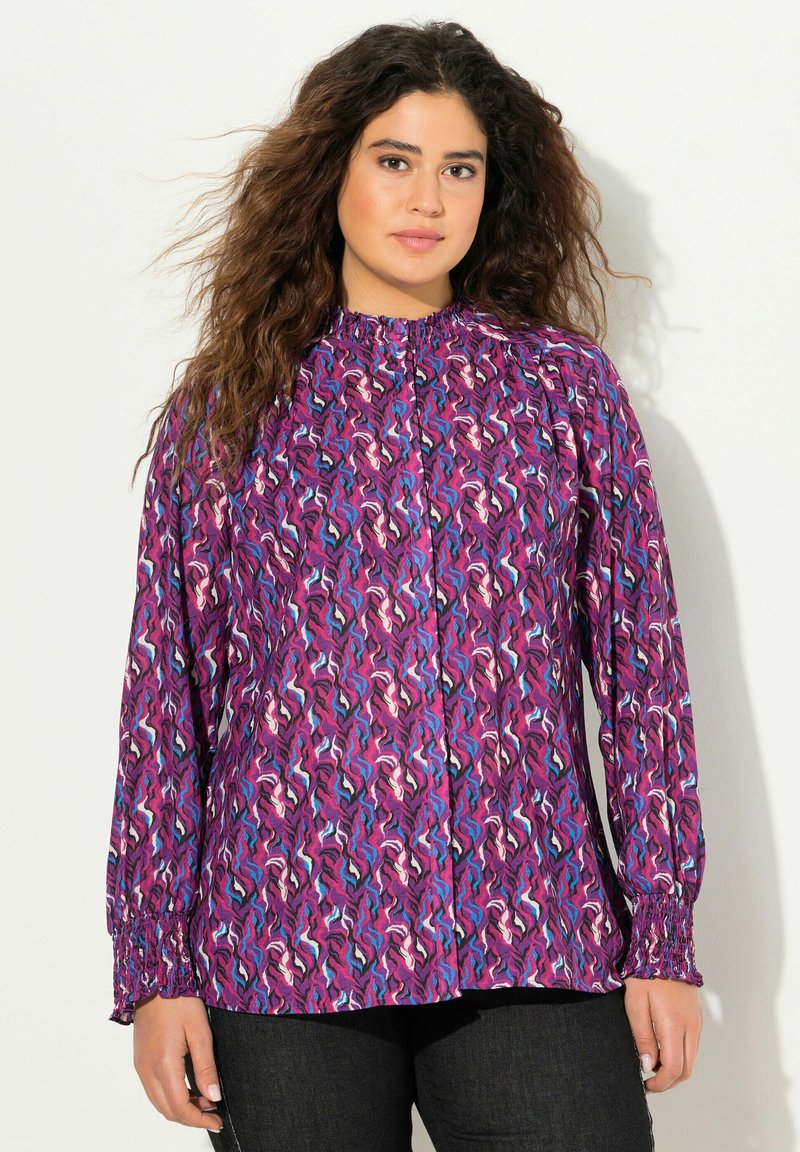 Blusa morada con diseño abstracto en espiral en tonos rosa, azul y blanco. Presenta un cuello alto y mangas largas con fruncidos y puños elásticos.
