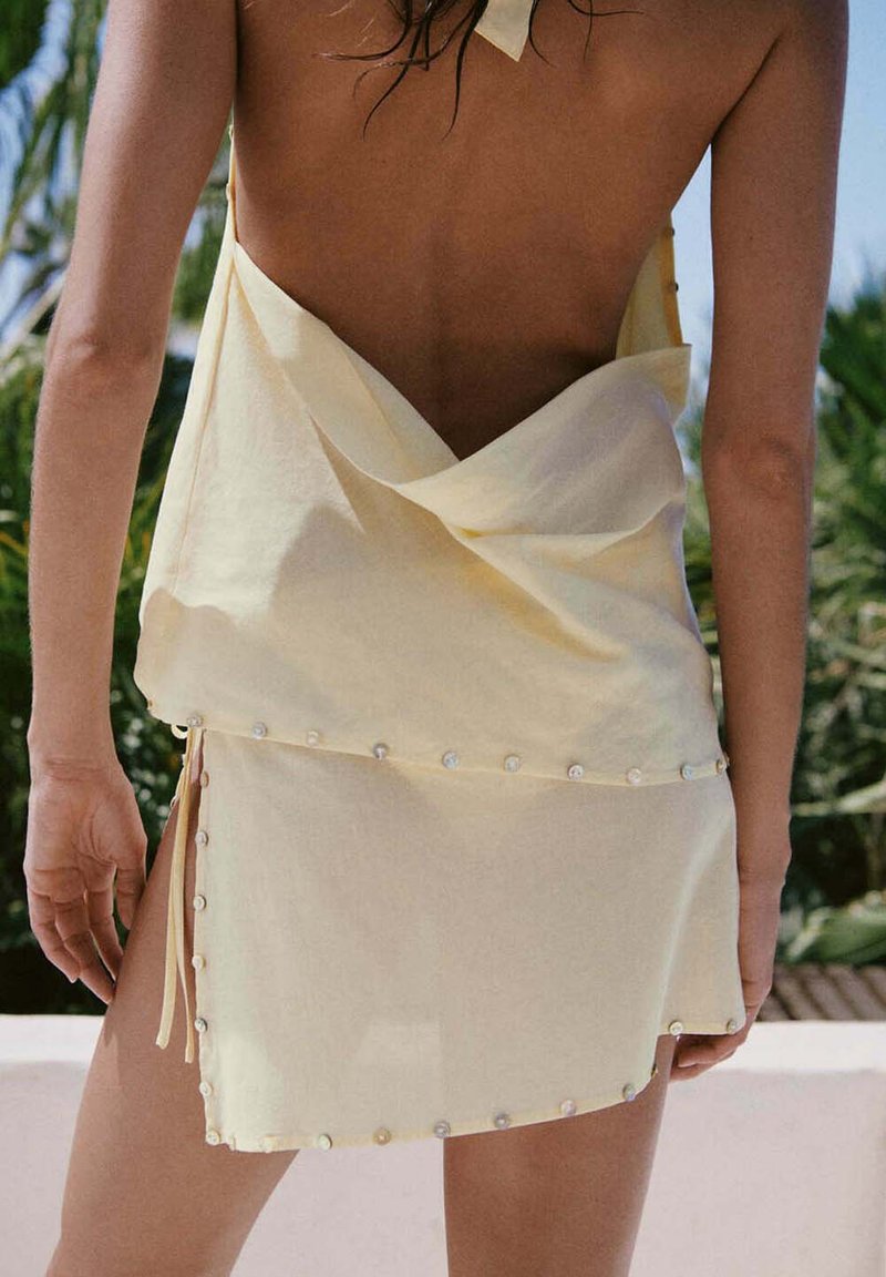 Femme portant une robe jaune clair dos nu avec des détails en perles, debout à l'extérieur devant un fond de verdure et de ciel dégagé.