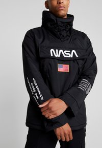 Zwarte pulloverjack met een capuchon, voorzien van een groot NASA-logo, een Amerikaanse vlagpatch en tekstdetails op de mouwen.