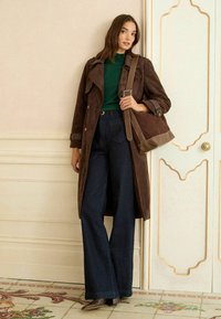 Hnědý semišový trench coat přes zelený top, spárovaný s širokými tmavými džínami a hnědými botami na podpatku; včetně hnědé kabelky přes rameno.