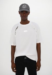 T-shirt blanche oversize avec des accents noirs et un patch logo, associé à une casquette grise avec un logo rond, le tout sur un fond neutre.