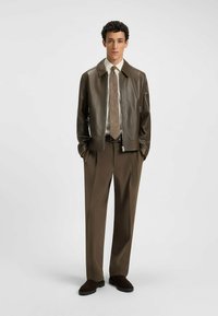 Brown leather jacket with a zip front, paired with a cream shirt and patterned tie. Loose-fitting brown trousers and dark footwear.