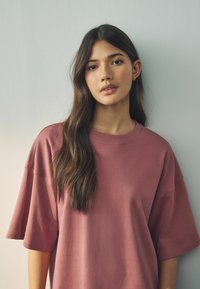 Preveliki kratkotrajni T-shirt u dusty rose boji s okruglim izrezom i glatkom teksturom. Model ima dugu, valovitu kosu.