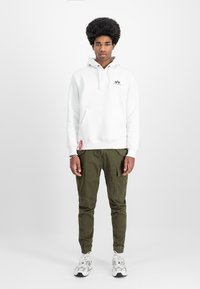 White hoodie with a front pocket and logo, paired with olive cargo pants featuring side pockets, and white sneakers with black accents.