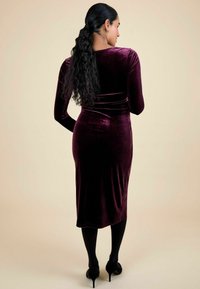 Rochie din catifea de un burgund profund, cu mâneci lungi și o siluetă ajustată, care ajunge până la jumătatea gambei, purtată cu colanți negri și tocuri.