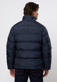 Marineblauwe pufferjack met een hoge kraag en horizontaal gestikte design. Beschikt over een gladde, glanzende textuur en elastische manchetten.