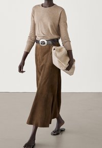 Femme portant un haut beige à manches longues, une jupe midi en suède marron, une ceinture noire avec une grande boucle argentée, tenant une pochette beige texturée.