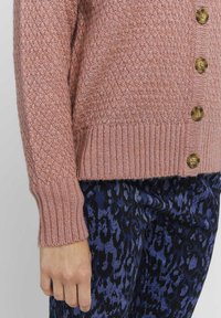 Cardigan tricoté dans une teinte rose douce avec un motif texturé, présentant un ourlet et des poignets côtelés, ainsi que des boutons dorés sur le devant.