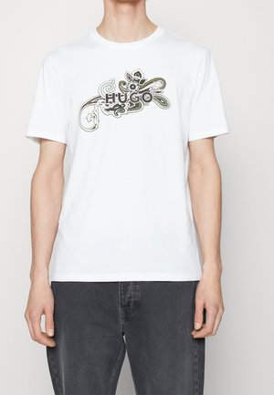 Man draagt een wit T-shirt met korte mouwen met een zwart-olijfgroen bloemenpatroon en de tekst "HUGO" over de borst, gecombineerd met donkergrijze broek.