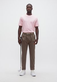 Άντρας που στέκεται, φοράει ροζ μπλουζάκι, καφέ παντελόνι, άσπρα αθλητικά παπούτσια, κρατώντας ένα γκολφ κλαμπ στο δεξί του χέρι πάνω σε λευκό φόντο.