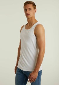 Top din bumbac alb, fără mâneci, cu decolteu rotund, având un logo subtil pe piept, asociat cu blugi ajustați de culoare albastră. Design simplu, într-o culoare solidă.