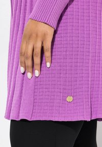 Lila Strickpullover mit strukturiertem Muster, gerippten Bündchen und einem goldenen Emblem am Saum. Schwarze Leggings sind teilweise darunter sichtbar.