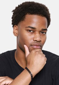 Bracciale a catena nera al polso, con maglie lucide. Il soggetto indossa una maglietta nera e posa in modo pensieroso con la mano sul mento. Il tono della pelle è medio.