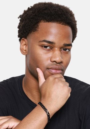 Bracelet en chaîne noire au poignet, avec des maillons brillants. Le sujet porte une chemise noire et pose pensivement la main sur le menton. Le teint est moyen.