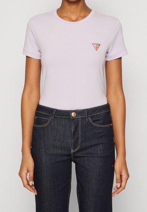 Camiseta de manga corta en color lila, hecha de material suave, con un pequeño logo triangular rojo en el lado izquierdo del pecho. Combinada con jeans de mezclilla oscura.