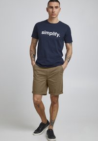 Tengerészkék póló, fehér "simplify." felirattal, világosbarna hajtott nadrággal és fekete tornacipővel párosítva. Tetovált karok láthatók.