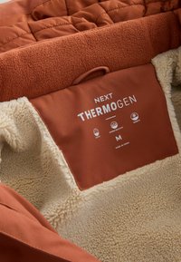 Interiorul jachetei termice este realizat dintr-un material textil quilted de culoare maro, cu o căptușeală moale din fleece crem, etichetă cu inscripția "NEXT THERMOGEN" și etichetă de mărime "M".