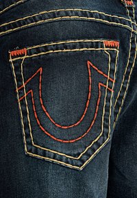 Los jeans de mezclilla oscura presentan un diseño distintivo en los bolsillos traseros con costuras naranjas que forman un patrón en forma de U, realzado por acentos amarillos.