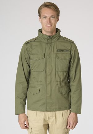 Giovane uomo sorridente, indossa una giacca verde oliva U.S. Polo Assn. con più tasche frontali e pantaloni beige, in piedi davanti a uno sfondo semplice.