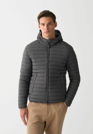 Chaqueta gris acolchada con capucha, que cuenta con un diseño de acolchado horizontal, cremallera frontal y bolsillos laterales. Textura suave y un detalle mínimo.