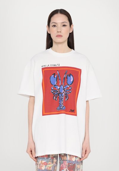 Tricou alb supradimensionat cu un grafic îndrăzneț de homar albastru într-un cadru pătrat roșu, cu textul "VIVE LA STIEGLITZ" deasupra.