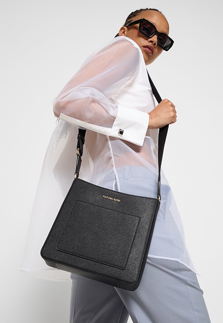 Schwarze strukturierte Schultertasche mit einer Fronttasche, goldenen Hardware-Akzenten und einem abnehmbaren Tragegurt, gehalten von einem Modell, das eine durchsichtige weiße Bluse trägt.