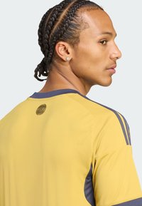 Gelbes Sporttrikot mit marineblauen Akzenten, rundem Kragen, strukturierten Schulterpaneelen und einem runden Logo auf der Rückseite in der Nähe des Kragens.