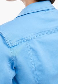 Hellblaue Jeansjacke mit klassischem Kragen, sichtbaren Nähten und einem glatten, strukturierten Stoff. Teilansicht von hinten.