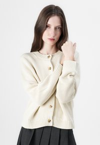 Kremfarget strikket cardigan med rund hals, utstyrt med treknapper og ribbestrikkede mansjetter, sammen med en mørk plisserte skjørt.