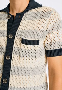 Kortärmad cardigan i krämfärg, grå och marinblå randig stickning. Har en texturerad, öppen väv, en ribbad krage och en bröstficka med kontrasterande kant.