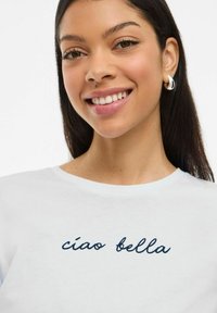 T-shirt blanc à encolure ronde, avec une broderie cursive bleu marine disant "ciao bella" sur le devant. Modèle portant des boucles d'oreilles en argent.