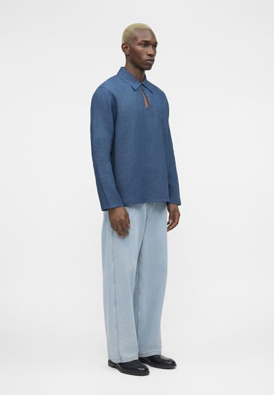 Άνδρας με κοντά ξανθά μαλλιά που φοράει μπλε μακρυμάνικο πουκάμισο με γιακά και άνοιγμα σε σχήμα κλειδιού, ανοιχτόχρωμα τζιν με φαρδιά μπατζάκια και μαύρα παπούτσια, όρθιος.