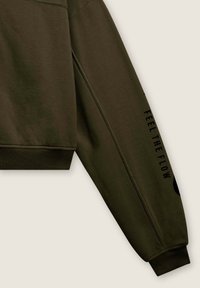 Manica di felpa verde oliva con polsino a costine, con un logo circolare nero e la scritta "FEEL THE FLOW" lungo il braccio. Texture liscia.