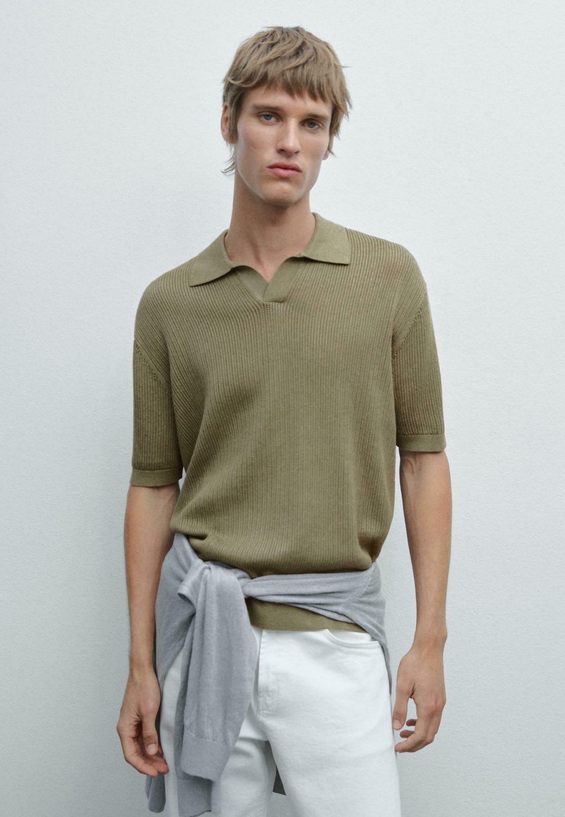 Polo shirt a righe verde oliva con colletto classico. Maglione grigio legato in vita. Pantaloni bianchi completano l'outfit.