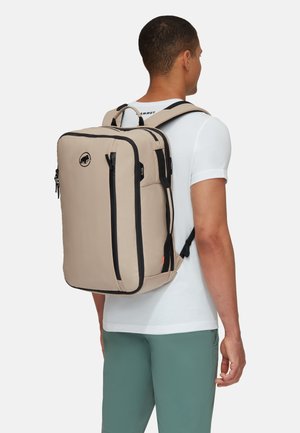 Beige rugzak met zwarte ritsen en accenten, rechthoekige vorm, gewatteerde schouderbanden en een logo ontwerp zichtbaar aan de voorkant.