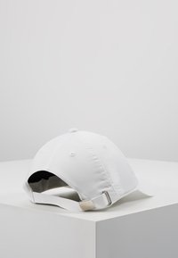 Fehér basebollap, sima, texturált anyagból. Állítható hátsó pántja van fém kiegészítőkkel és lekerekített napellenzővel.