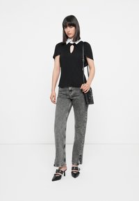 Schwarzes Oberteil mit einem weißen Kragen und einem Schlüsselloch-Detail, kombiniert mit grauen Jeans mit fransenbesäumten Säumen und schwarzen, mit Riemenverzierungen versehenen High Heels.