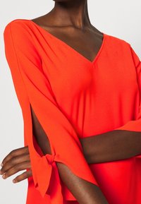 Blouse orange à col en V avec manches larges, fentes latérales et nœuds aux poignets, en tissu lisse et léger.