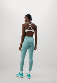 Person med fletninger iført en hvid sports-bh, lyseblå højtsiddende leggings, hvide sokker og turkise sneakers, som står foran en ensfarvet baggrund.