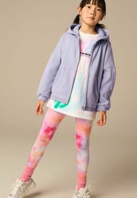 Světle fialová mikina na zip, bílý tričko s tie-dye akcenty, vícebarevné legíny s růžovými manžetami a bílé tenisky.