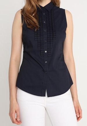 Sleeveless navy blue button-up blouse with decorative pleated front panels worn with white pants.