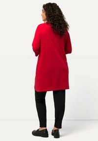 Rote Langarm-Tunika aus weichem Stoff, kombiniert mit schwarzen Slim-Fit-Hosen, getragen mit schwarzen Schuhen, von hinten gesehen.