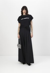 Melnā T-kreklā ar "KARL LAGERFELD" logo, kombinācijā ar melnām platajām biksēm. Aksesuāri ietver mazu sudraba rokassomu. Minimalistisks dizains.