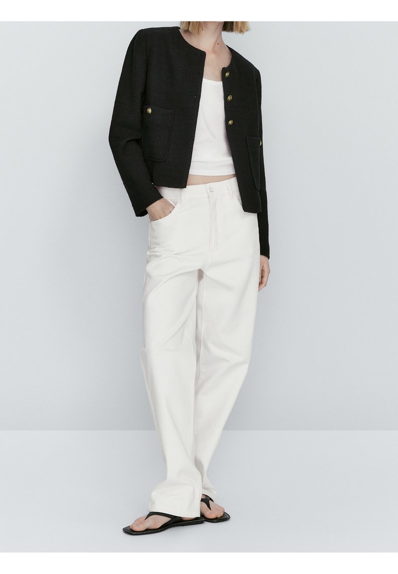 Svart kort jacka med guldknappar och två framfickor, matchad med vita vida byxor och svarta flip-flops.