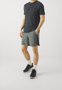 Muž v černém tričku Under Armour, šedých sportovních šortkách, bílých ponožkách a černých běžeckých botách, stojí s jednou rukou v kapse.
