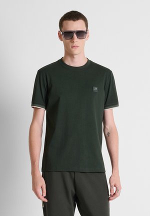Giovane uomo che indossa grandi occhiali da sole scuri e una maglietta a maniche corte verde scuro con un piccolo logo sul petto, in piedi davanti a uno sfondo semplice.