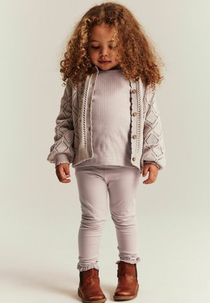 Małe dziecko z kręconymi włosami, ubrane w beżowy dziergany kardigan, jasną bluzkę i legginsy oraz brązowe botki za kostkę, stojące na tle o neutralnym kolorze.